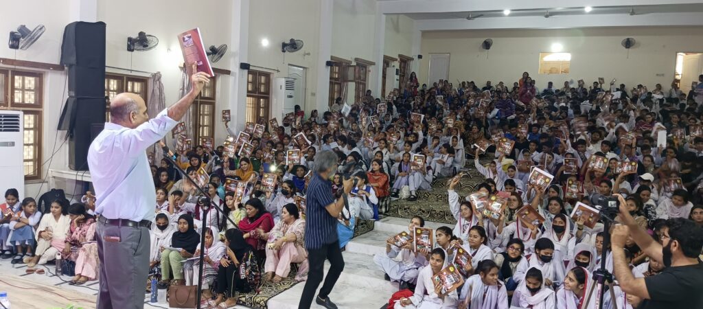 A wide shot captures a large indoor gathering of over 650 students, mostly female, seated on the floor and in chairs, facing a man standing on a slightly raised platform at the left. The man, identified as Anser Javed, Executive Director of Chopan Trust, is holding a book aloft with his right hand and appears to be speaking. He is dressed in a light blue shirt and dark trousers. To his right, another man with a camera seems to be taking pictures of the audience. The students are holding up similar books, many smiling and looking towards the speaker. A large black speaker stands behind Anser Javed. The room is brightly lit with fluorescent lights overhead and has light-colored walls with several windows and doorways visible in the background.