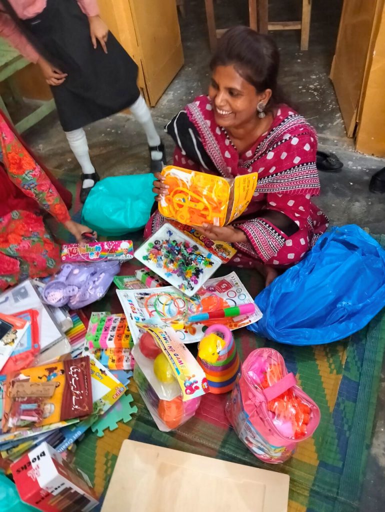 A smiling woman in a pink dress holds up a new educational toy, surrounded by many colorful learning aids and toys on the floor.